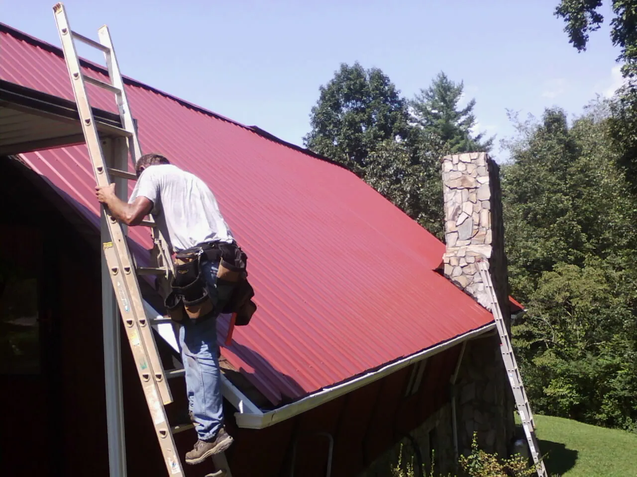 Roofer on a ladder beside a red metal roof and stone chimney — Soffit & Fascia Repair by Roofing Brothers in Schaefferstown